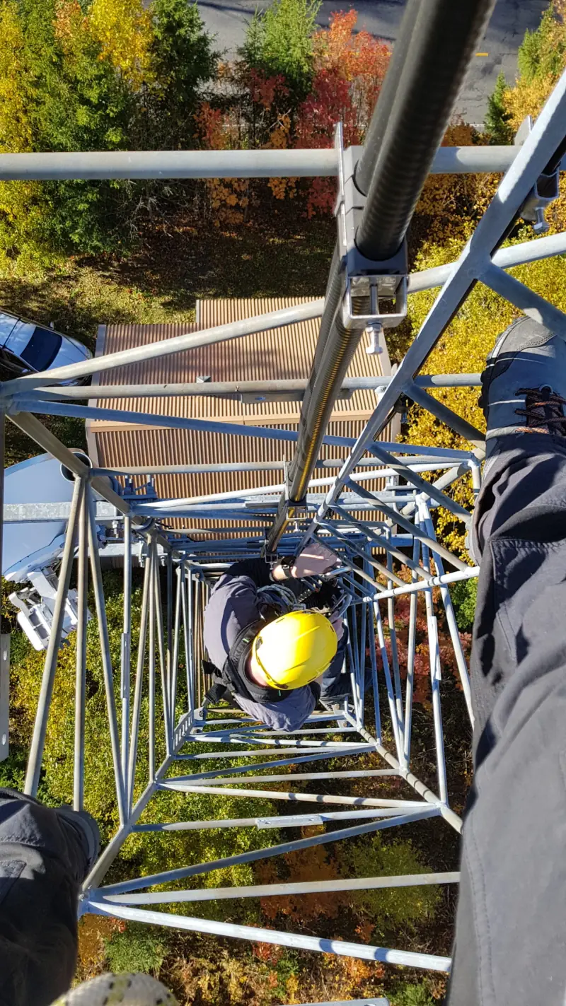 Looking down a telecoms mast at a climber in a yellow helmet, with autumn trees below